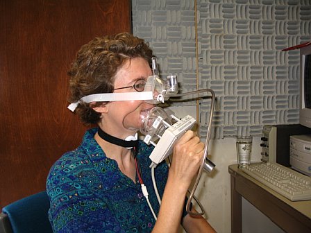 Photo of Natasha Warner in a blue-green shirt with glasses and short curly brown hair, from the side, holding an oral airflow mask over her mouth, a nasal airflow mask strapped over the nose with velcro around the back of the head, and EGG sensors strapped around the neck with velcro. The equipment has cables/tubes attached to it.  Background shows soundproofing on a wall and the corner of a table.